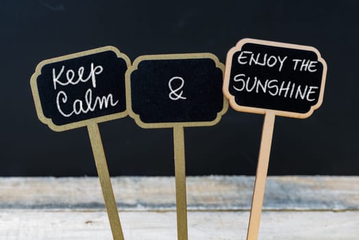 Keep Calm and Enjoy The Sunshine message written with chalk on mini blackboard labels, defocused chalkboard and wooden table in background. Fun and humor concept