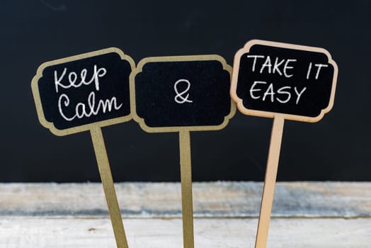 Keep Calm and Take It Easy message written with chalk on mini blackboard labels, defocused chalkboard and wooden table in background. Fun and humor concept