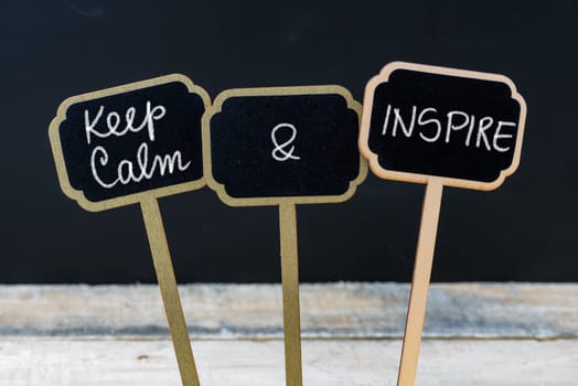 Keep Calm and Inspire message written with chalk on mini blackboard labels, defocused chalkboard and wooden table in background. Fun and humor concept