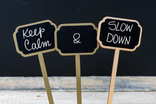 Keep Calm and Slow Down message written with chalk on mini blackboard labels, defocused chalkboard and wooden table in background. Fun and humor concept