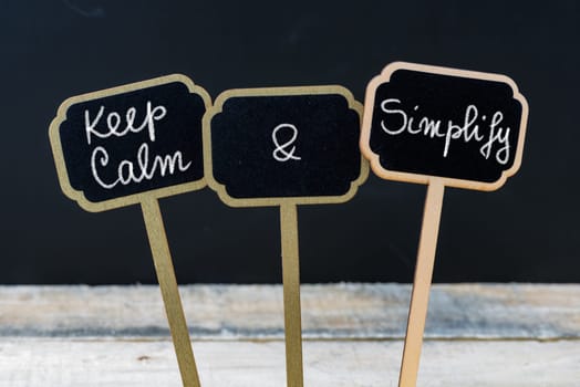 Keep Calm and Simplify message written with chalk on mini blackboard labels, defocused chalkboard and wooden table in background. Fun and humor concept