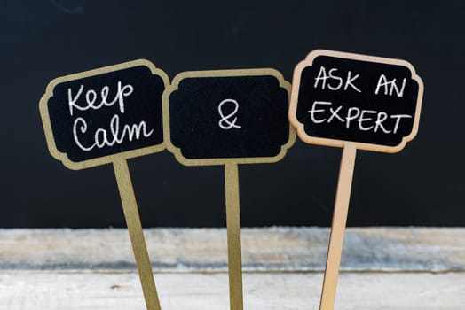 Keep Calm and Ask An Expert message written with chalk on mini blackboard labels, defocused chalkboard and wooden table in background. Fun and humor concept