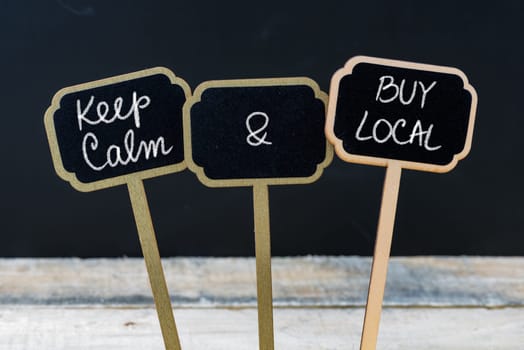 Keep Calm and Buy Local message written with chalk on mini blackboard labels, defocused chalkboard and wooden table in background. Fun and humor concept