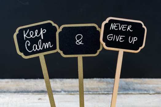 Keep Calm and Never Give Up message written with chalk on mini blackboard labels, defocused chalkboard and wooden table in background. Fun and humor concept