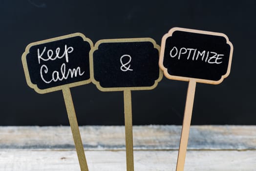 Keep Calm and Optimize message written with chalk on mini blackboard labels, defocused chalkboard and wooden table in background. Fun and humor concept