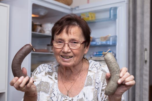 Happy senior woman holding pork liver sausages while standing in front of the open fridge in the kitchen.