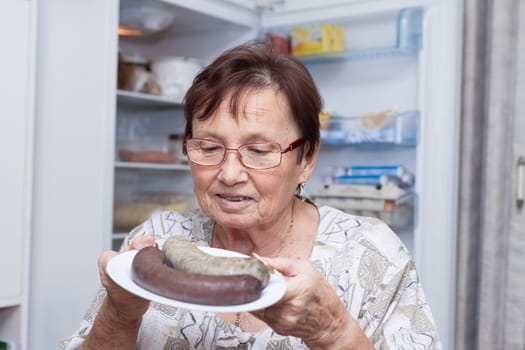 Happy senior woman holding a plate with pork liver sausages while standing in front of the open fridge in the kitchen.