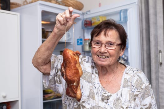 Happy senior woman holding smoked meat while standing in front of the open fridge in the kitchen.