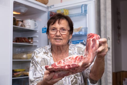 Happy senior woman holding raw pork ribs while standing in front of the open fridge in the kitchen.