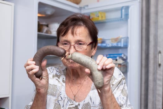 Senior woman holding pork liver sausages while standing in front of the open fridge in the kitchen.