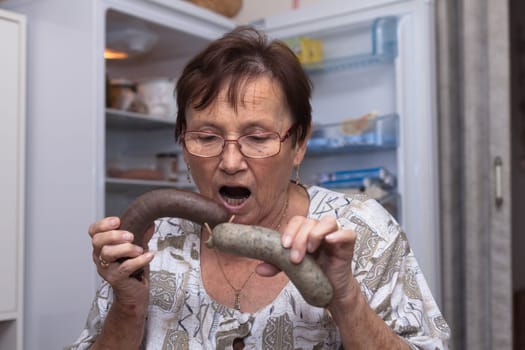 Surprised senior woman holding pork liver sausages while standing in front of the open fridge in the kitchen.