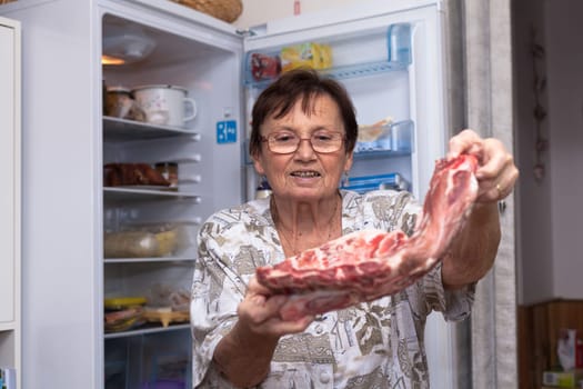 Senior woman holding raw pork ribs while standing in front of the open fridge in the kitchen.