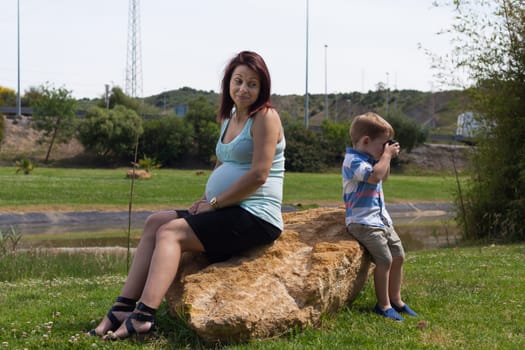 Pregnant woman and child boy sitting back to back outdoors.