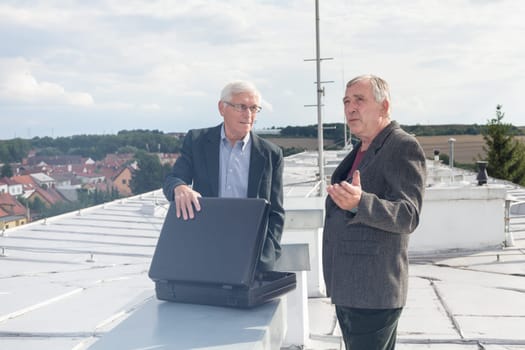 Two senior businessmen with briefcase discussing business deal outdoors on the roof of a building.