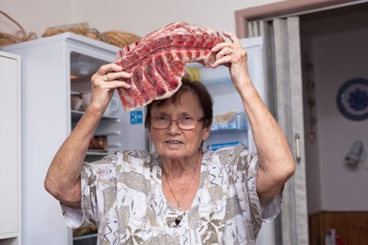 Senior woman holding raw pork ribs while standing in front of the open fridge in the kitchen.