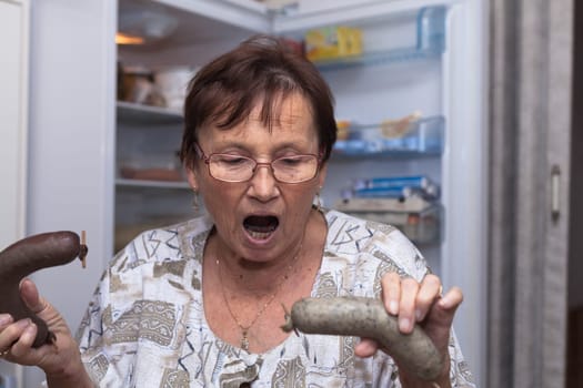 Shocked senior woman holding pork liver sausages while standing in front of the open fridge in the kitchen.
