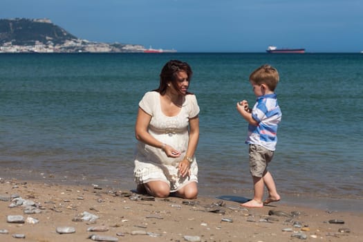 Happy pregnant woman playing with child boy and enjoying sunny day on the beach in Spain.