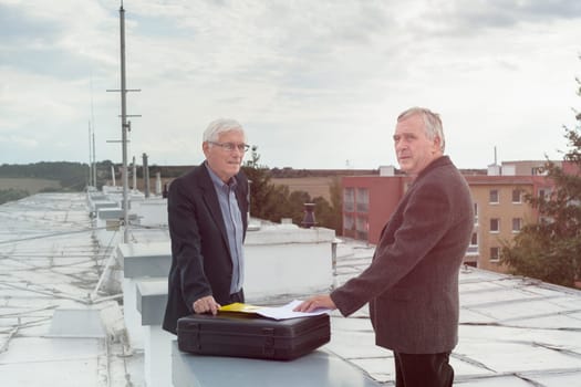 Two senior businessmen with documents making a business deal outdoors on the roof of a building.