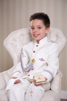 Young boy with prayer book and rosary celebrating his First Holy Communion sitting on an armchair and looking at camera.