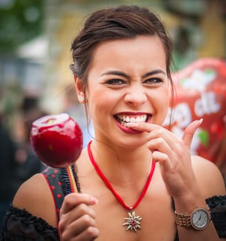 Attractive young woman in traditional Dirndl dress holding a red caramelized apple at the Oktoberfest with people in the background.