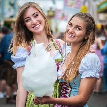 Two young women in traditional Bavarian clothes, dirndl or tracht, smiling with cotton candy floss at the Oktoberfest