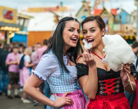 Two young women in traditional Bavarian clothes, dirndl or tracht, laughing with cotton candy floss at the Oktoberfest
