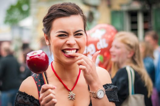 Attractive young woman in traditional Dirndl dress holding a red caramelized apple at the Oktoberfest with people in the background.