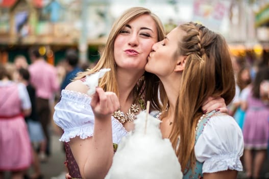 Two young women in traditional Bavarian clothes, dirndl or tracht, kissing with cotton candy floss at the Oktoberfest