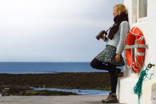 Young Caucasian blonde woman holding binoculars by seaside near a lighthouse with a life buoy