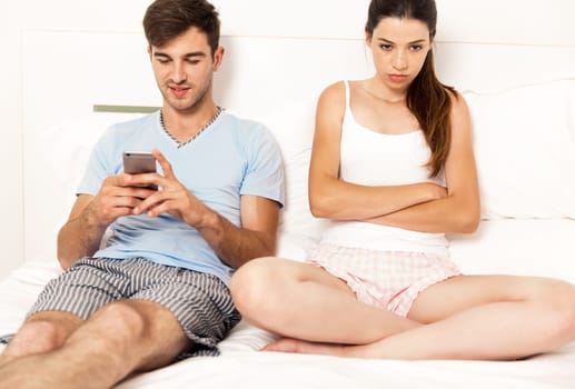 Addicted young man on bed texting while woman looks angry