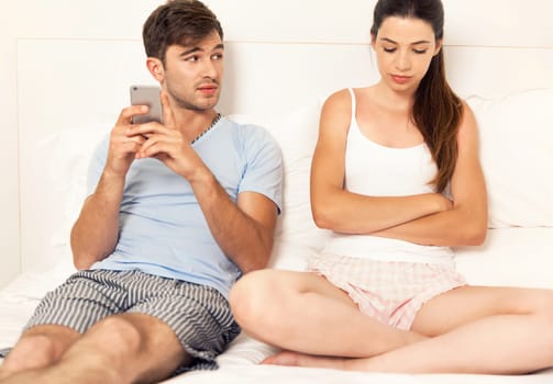 Addicted young man on bed texting while woman looks angry