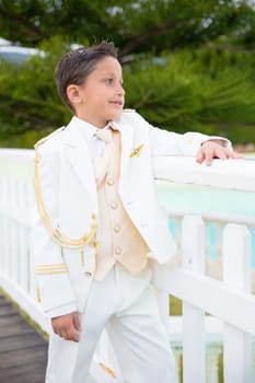 Young boy with white sailor suit leaning on a white wooden fence in his First Communion
