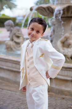 Young boy with white sailor suit smiling in his First Communion standing in front of a fountain
