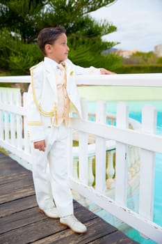 Young boy with white sailor suit leaning on a white wooden fence over a bridge in his First Communion