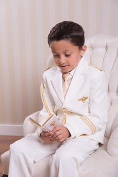 Young boy sitting on an armchair and reading a prayer book in his First Holy Communion.