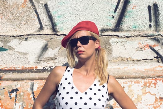 Horizontal portrait of woman with polka dress, red hat and black sun glasses against a graffiti wall