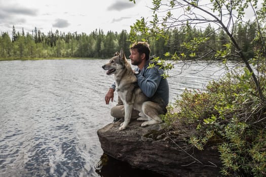 Handsome man and dog are sitting on the stone near of lake and looking into the distance