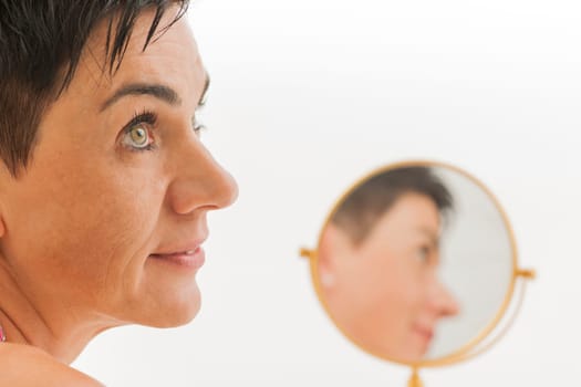 Closeup of a middle-aged black-haired woman with a round make-up mirror against a white background looking to the side.