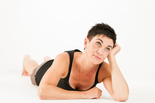 Full-body view of a black-haired middle-aged woman barefoot in shorts and black top lying on her stomach against a white background looking dreamily upwards.