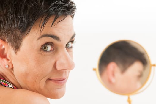 Closeup of a middle-aged black-haired woman with a round make-up mirror against a white background looking into the camera.
