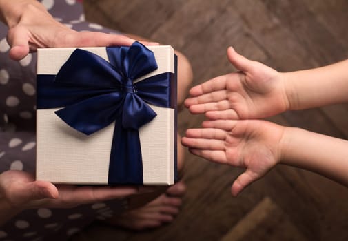 Top view of hands of mother giving one large gift box with blue ribbon to her kid with open hand