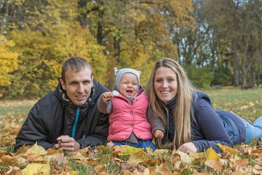 Smiling young parents with their laughing baby daughter are lying on the ground and looking at the camera in a beautiful autumn park