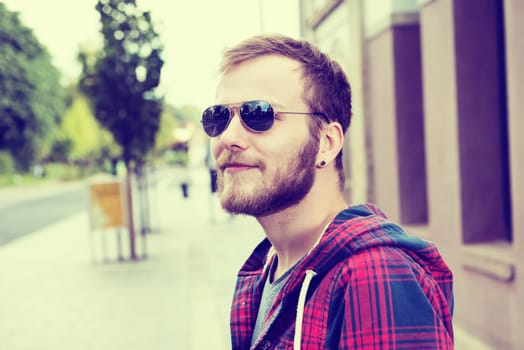 young bearded man with sunglasses in checkered shirt standing in front of city background