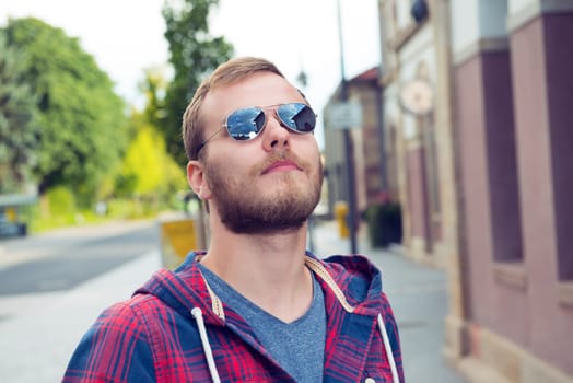 young bearded man with sunglasses in checkered shirt standing in front of city background