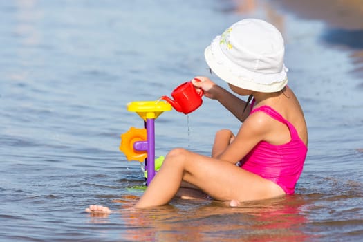 Five-year girl in a pink bathing suit sitting in on the river bank and plays in the sand toys