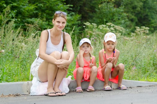 Mother and two children sitting on the curb of the road waiting for the bus