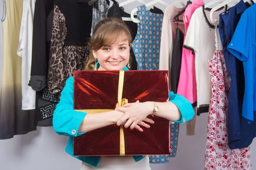 Happy young girl standing with big red gift on background of clothes on hangers