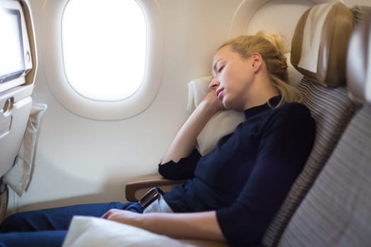 Tired blonde casual caucasian lady napping on seat while traveling by airplane. Commercial transportation by planes.