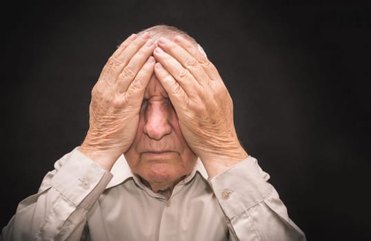 Portrait of an elderly man with face closed by hands on black with copyspace