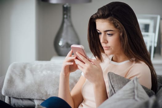 Teenage girl sitting on the sofa in her home, using a smart phone.